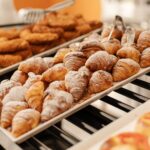 A selection of various baked pastries is arranged neatly on a tray in a bakery. The warm croissants with powdered sugar are highlighted, enticing customers.