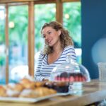 Female customer leaning on wooden counter at cafe with cupcakes under glass dome and croissants. Artisan, bakery, cozy, inviting, cheerful, rustic, premium